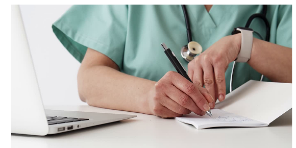 The hands of a medical professional in scrubs writing notes on a notebook.